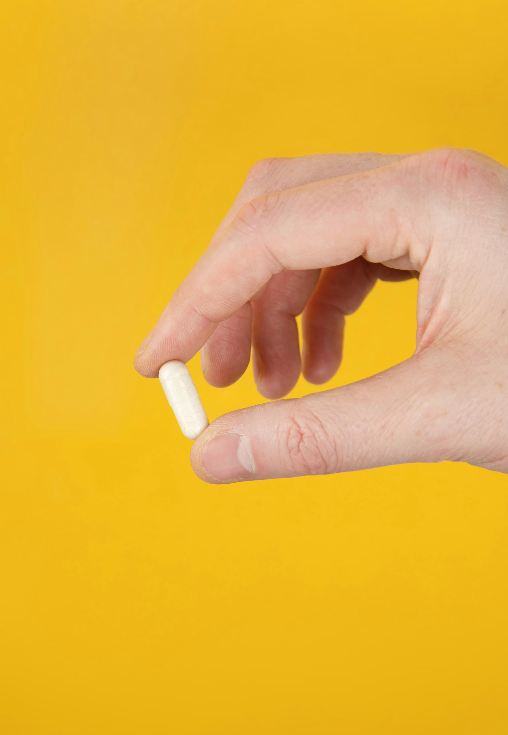 Hand holding a single white capsule between fingers against a bright yellow background.