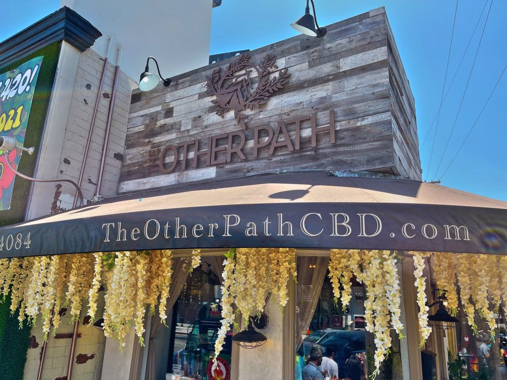 Exterior of The Other Path CBD storefront featuring a rustic wood sign, floral awning décor, and the website TheOtherPathCBD.com displayed above the entrance.