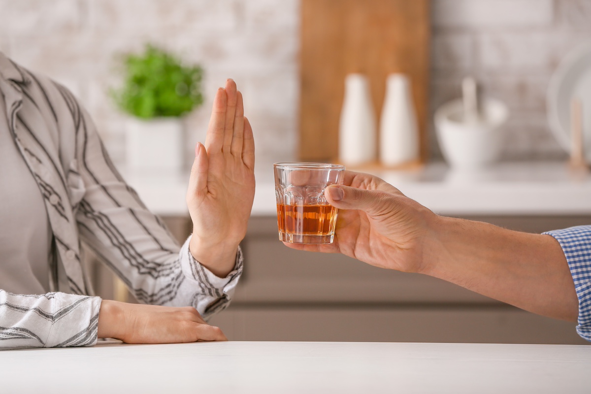 Person raising a hand to refuse a glass of alcohol being offered across a table in a kitchen setting.