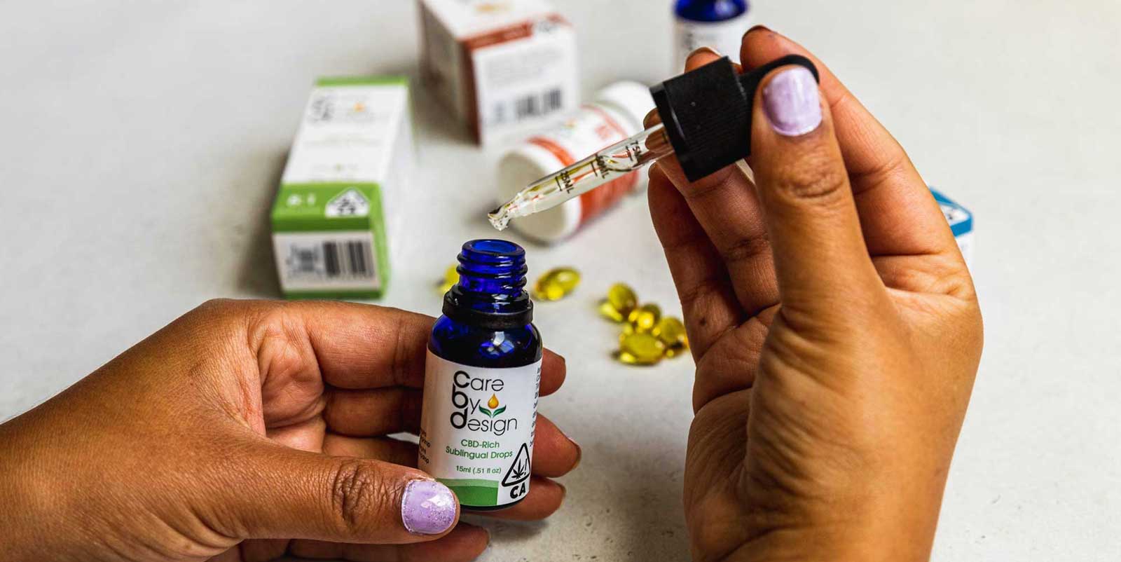 Hands holding a CBD sublingual oil bottle and dropper, dispensing CBD oil, with supplement capsules and packaging in the backgroun