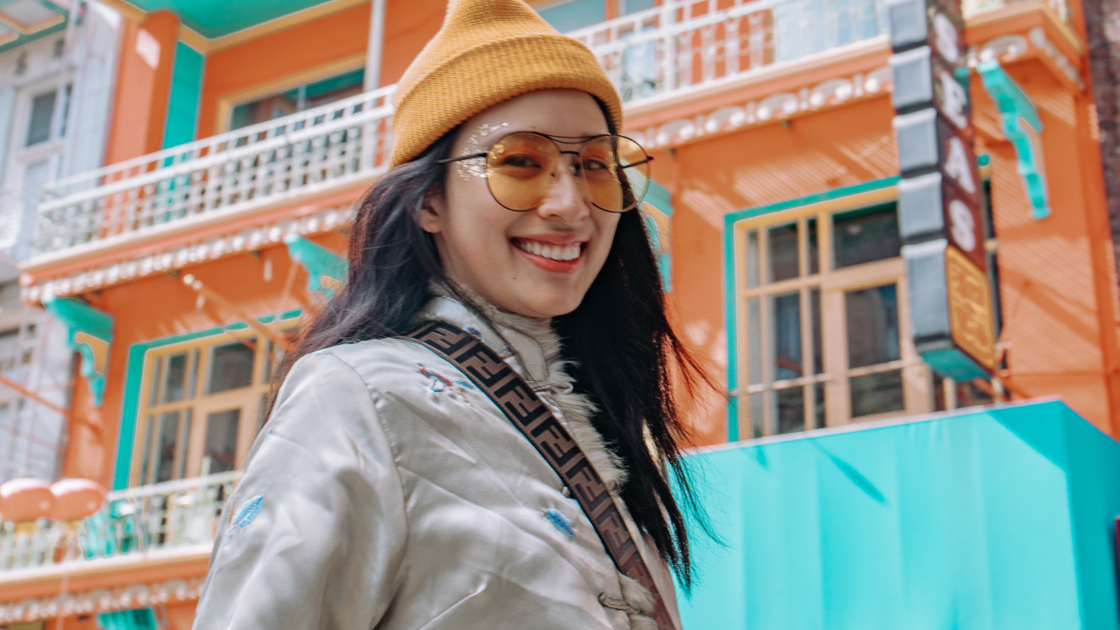 Smiling woman wearing a mustard beanie and round sunglasses walking past a colorful orange and teal building outdoors.