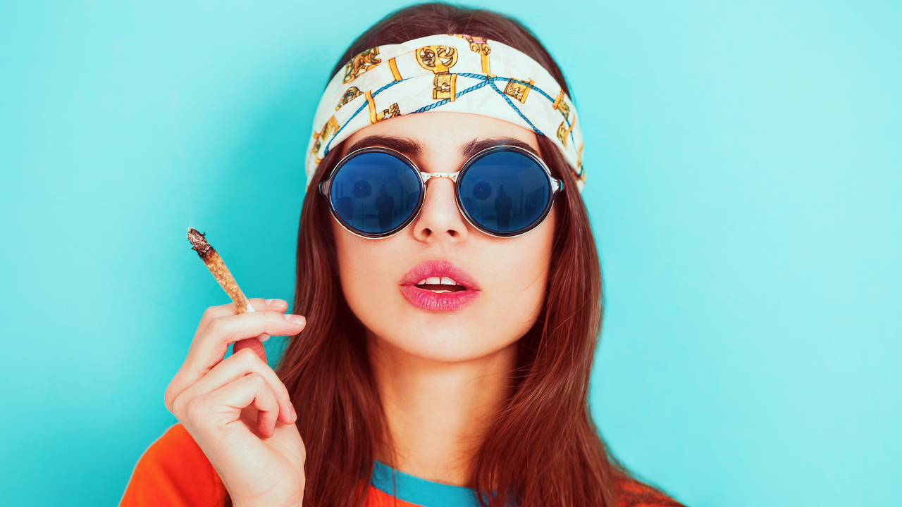 Woman wearing round sunglasses and a patterned headband holding a lit cannabis joint against a turquoise background.