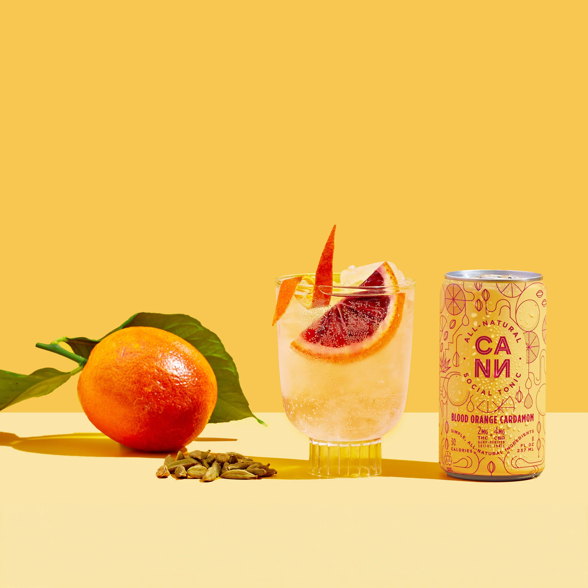 Blood orange and cardamom CANNA cannabis-infused social tonic shown with a garnished sparkling drink, fresh orange, and cardamom pods on a yellow background.