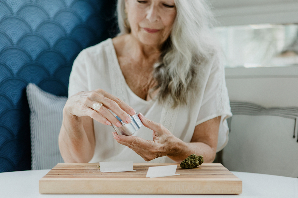 Older woman preparing medical cannabis, holding a small metal container over a wooden tray with rolling papers and a cannabis bud at home.