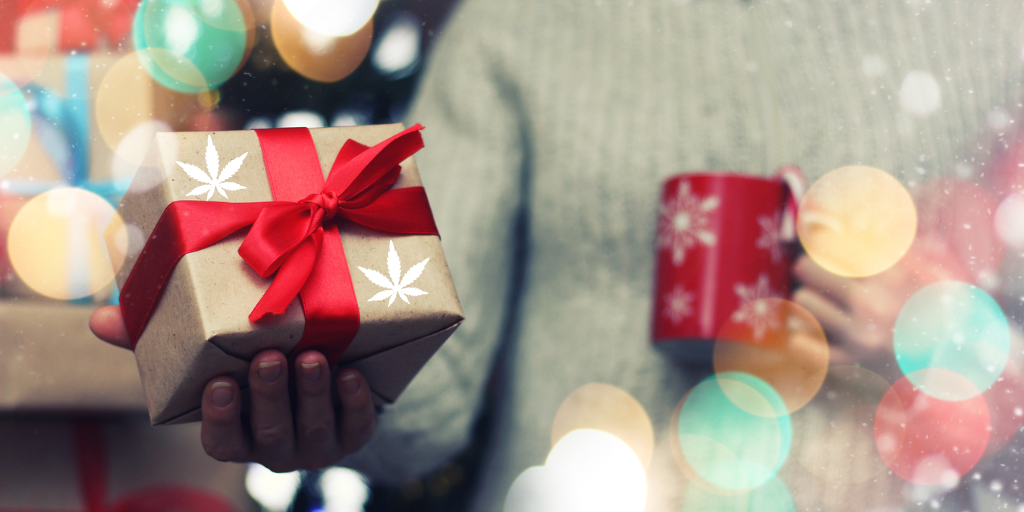 Person holding a gift wrapped in brown paper with a red ribbon and white cannabis leaf icons, while holding a festive red mug, with soft holiday lights in the background.
