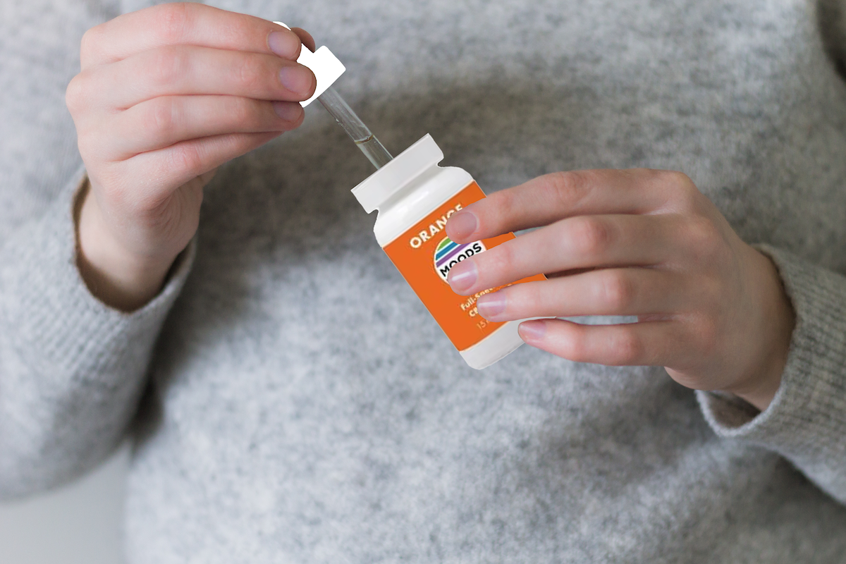 Hands holding a small orange-labeled bottle and dropper, dispensing liquid from a wellness oil supplement container.