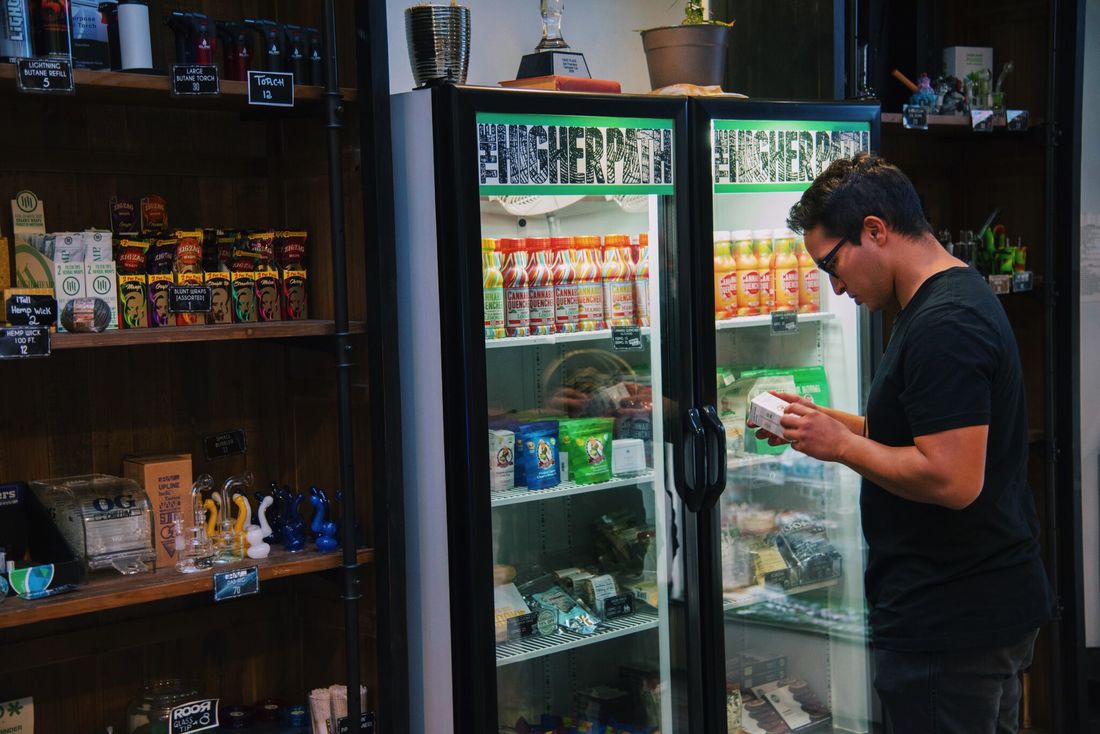 Customer browsing cannabis products in a dispensary, reading a product label beside a refrigerated display of edibles and beverages.