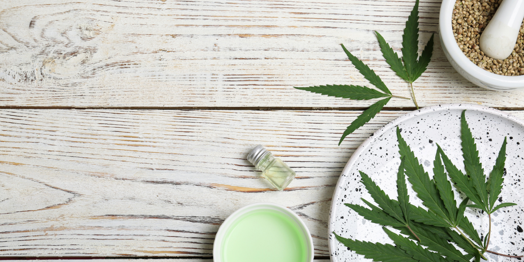 Flat lay of cannabis leaves with a small bottle of oil, green cream jar, and hemp seeds arranged on a white wooden surface.