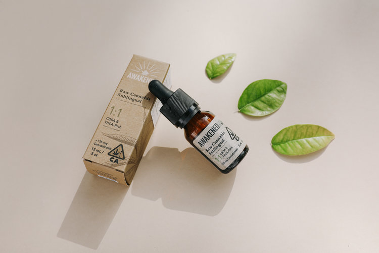 A brown glass dropper bottle and cardboard packaging for Awakened Raw Cannabis Sublingual 1:1 tincture, positioned next to three green leaves on a light-colored surface.