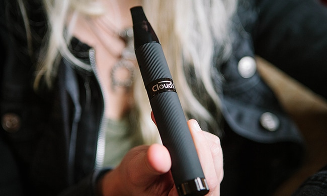 Close-up of a person holding a black vape pen with ‘Cloud’ branding, with the background softly out of focus.