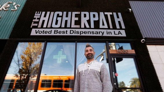 Exterior of The Higher Path dispensary with a sign reading ‘The Higher Path – Voted Best Dispensary in LA,’ showing a man standing in front of the entrance windows.