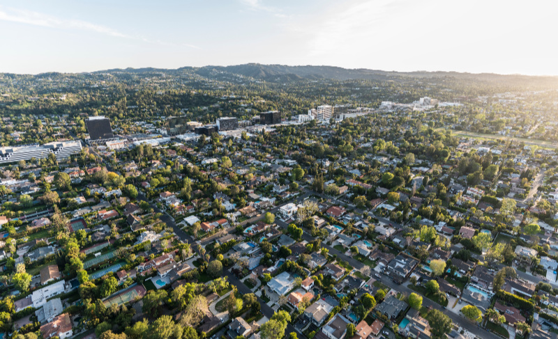 Aerial view of a leafy suburban neighborhood with tree-lined streets, low-rise buildings, and rolling hills in the background during golden-hour light.