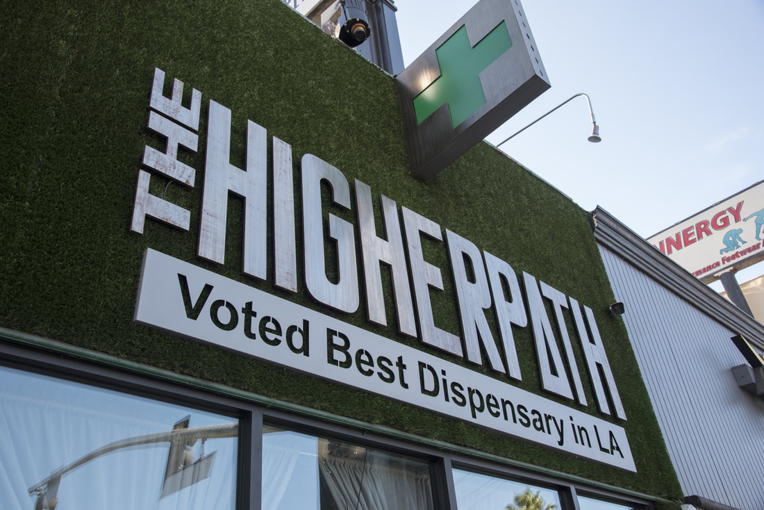 Exterior storefront sign reading ‘The Higher Path’ with a green cross above and a banner stating ‘Voted Best Dispensary in LA.’