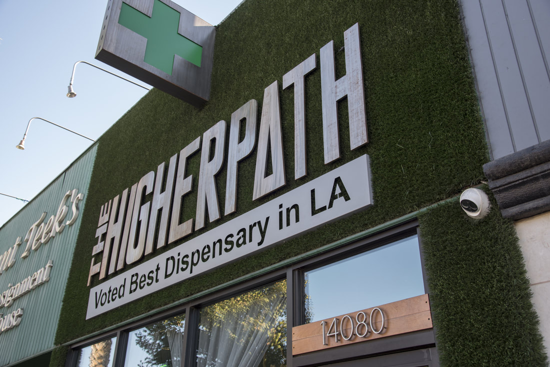 Exterior of The Higher Path cannabis dispensary in Los Angeles, featuring a green cross sign and a storefront sign reading ‘The Higher Path – Voted Best Dispensary in LA.’