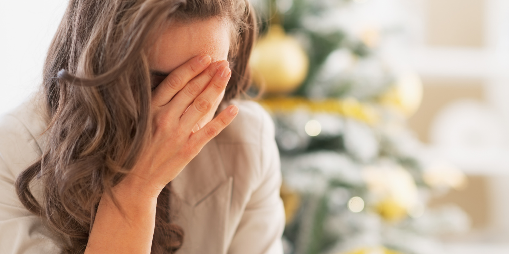 Woman covering her face in distress, sitting indoors near a decorated Christmas tree.