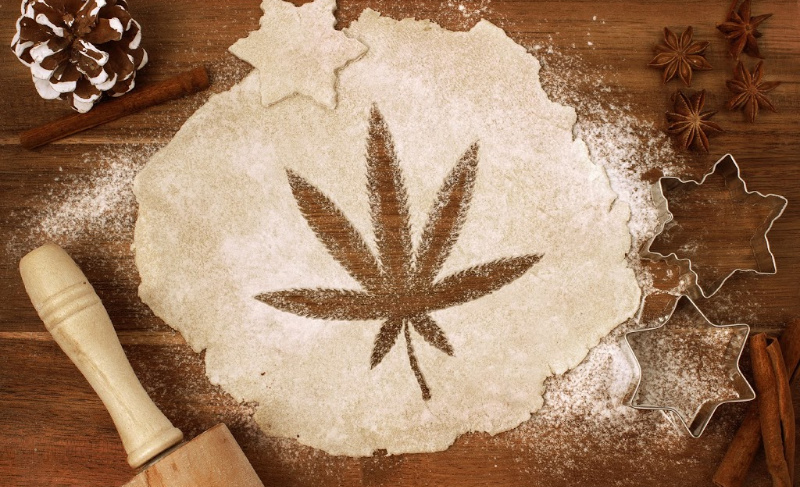 Rolled-out cookie dough dusted with flour featuring a cannabis leaf imprint, surrounded by baking tools, star-shaped cutters, cinnamon sticks, and spices on a wooden surface.