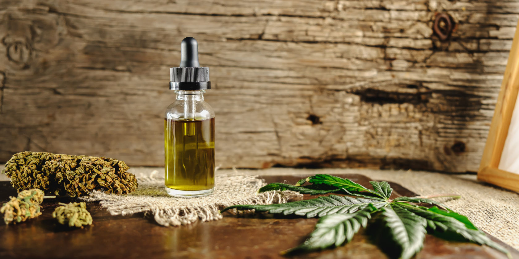 Glass dropper bottle of cannabis oil displayed with marijuana buds and leaves on a rustic wooden surface.