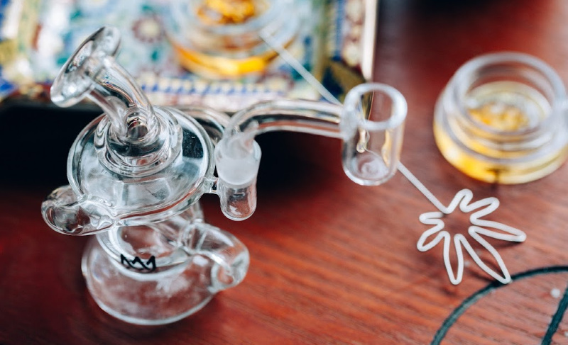 Glass dab rig with a quartz banger and metal dab tool resting on a wooden surface, with cannabis concentrate jars in the background.