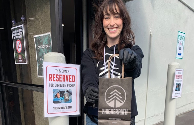 A smiling employee in black gloves holds out a The Higher Path shopping bag in front of a storefront with signs for reserved curbside pickup.