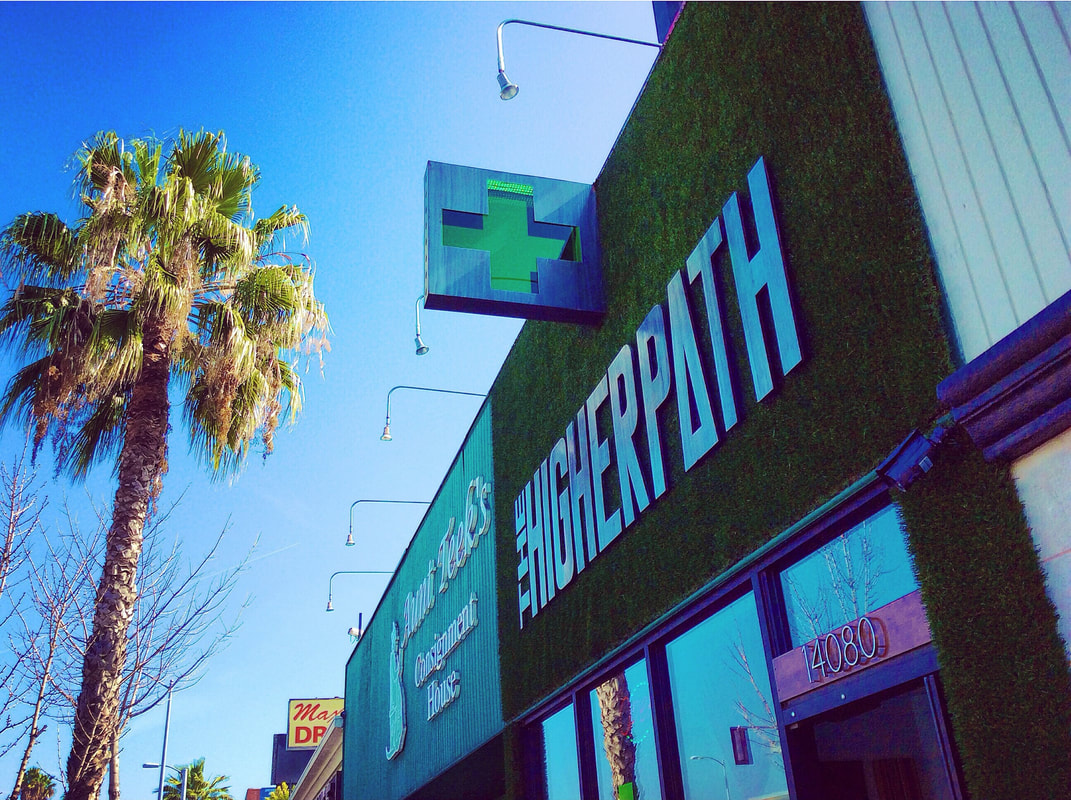Exterior of The Higher Path cannabis dispensary with green signage and cross logo on a sunny street lined with palm trees.