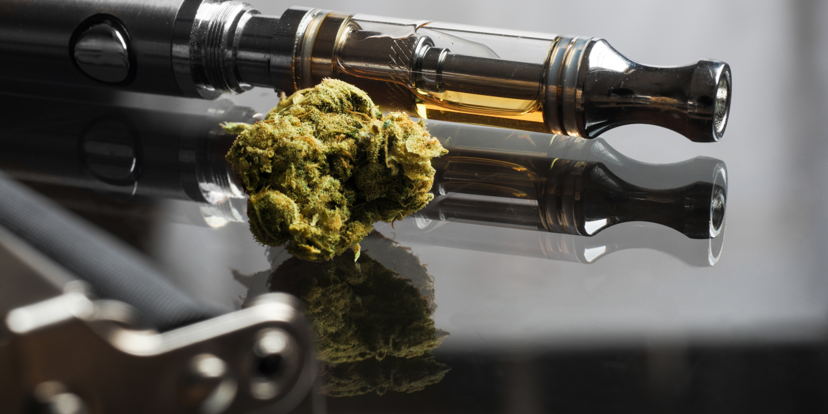 Close-up of a vape pen filled with cannabis oil resting beside a cannabis bud on a reflective surface.
