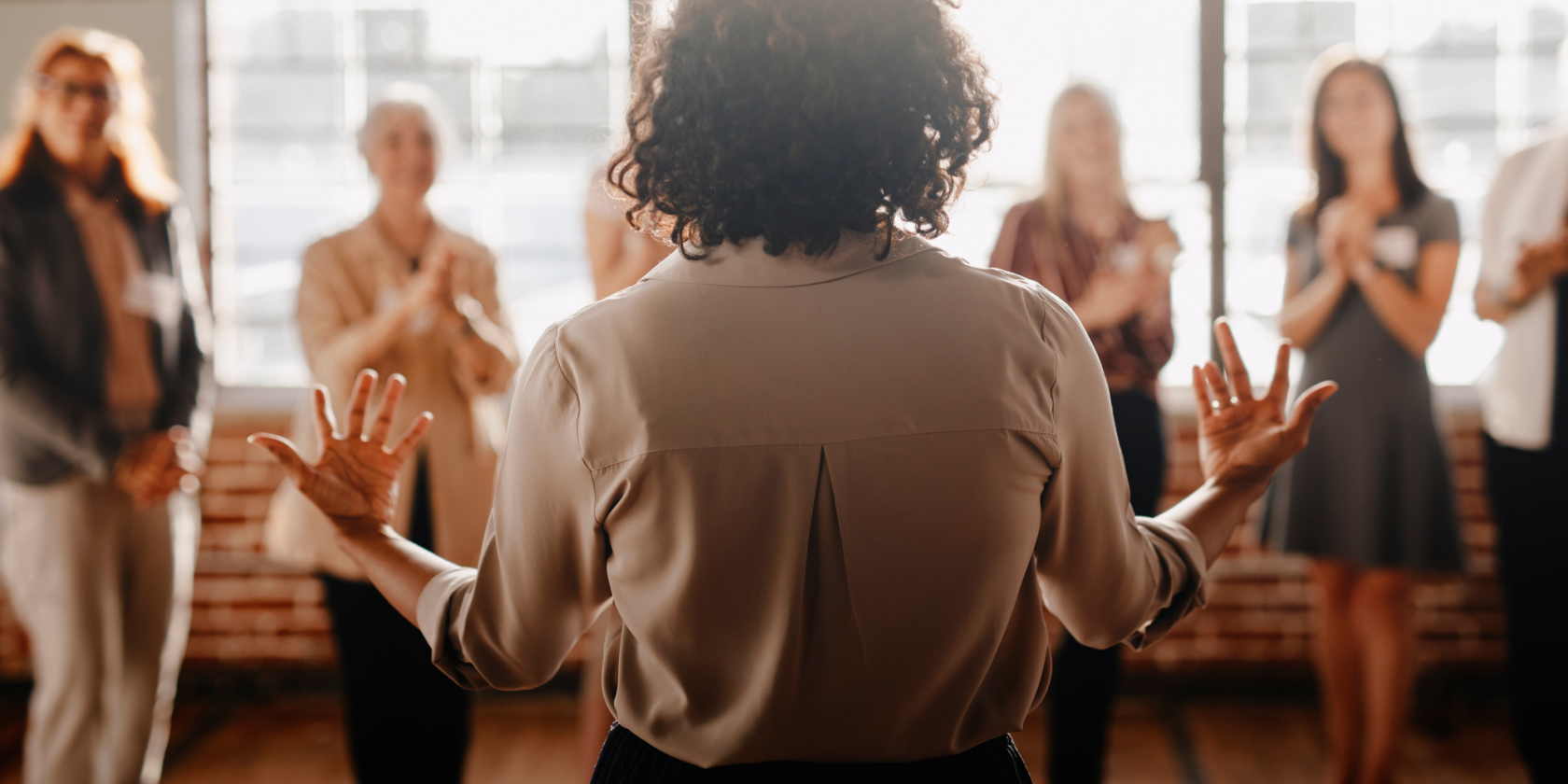 Person standing with hands raised while addressing a small group of people who are applauding in a bright, indoor meeting space.